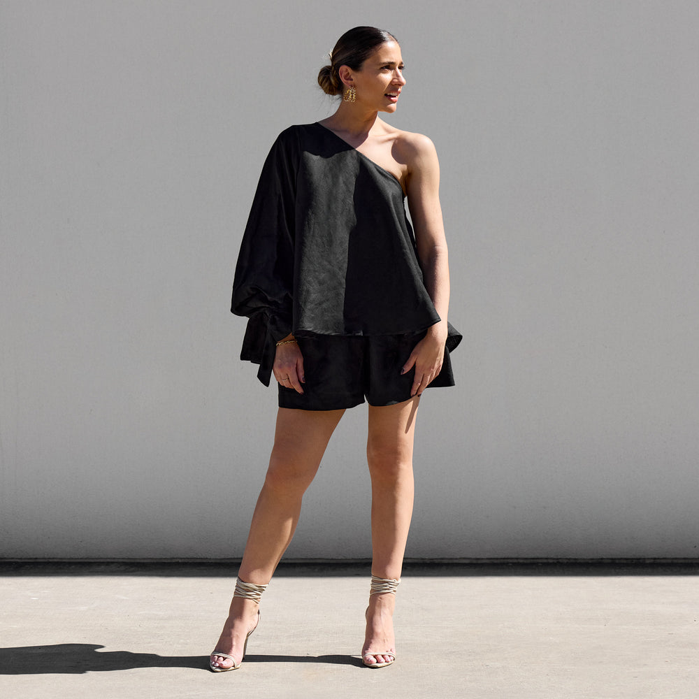 
                      
                        In front of a plain gray wall, a person with short hair wears Isabella Longginous Black OS Balloon Sleeve Swing Top paired with sandals. Facing slightly to the side in a relaxed pose, the sunny outdoor setting highlights their effortlessly chic style.
                      
                    