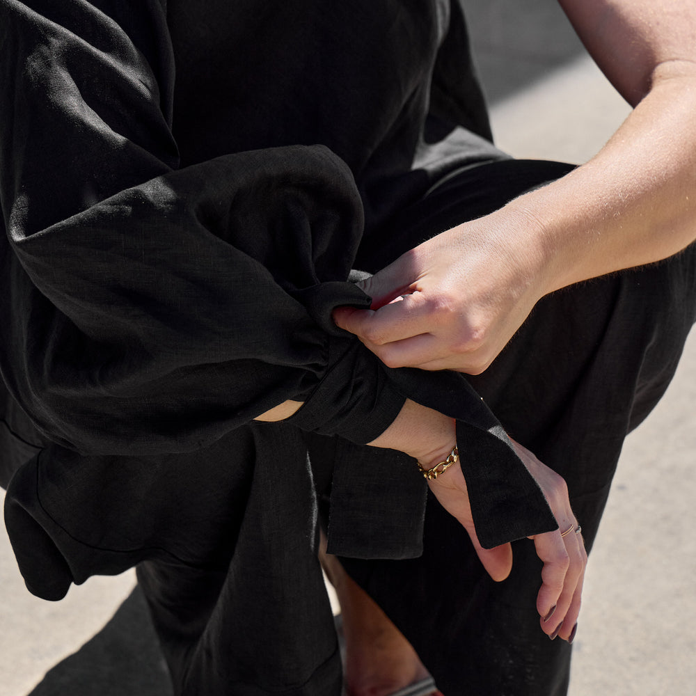 
                      
                        A person kneels on the concrete, adjusting the knot on an Isabella Longginou Black OS Balloon Sleeve Swing Top. They wear a gold bracelet on their right wrist and white open-toe sandals, with sunlight casting shadows around them.
                      
                    