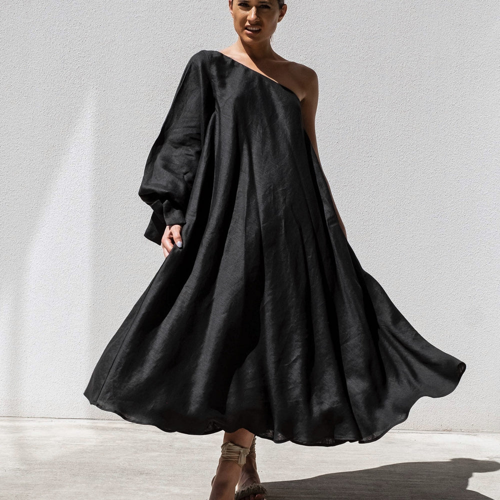 
                      
                        Wearing Isabella Longginous Black Balloon Sleeve Swing Dress, a person stands elegantly against a white wall. The Australian-designed one-shoulder dress features flowing fabric and stylish balloon sleeves. Strappy sandals complete the look as lighting casts a shadow.
                      
                    