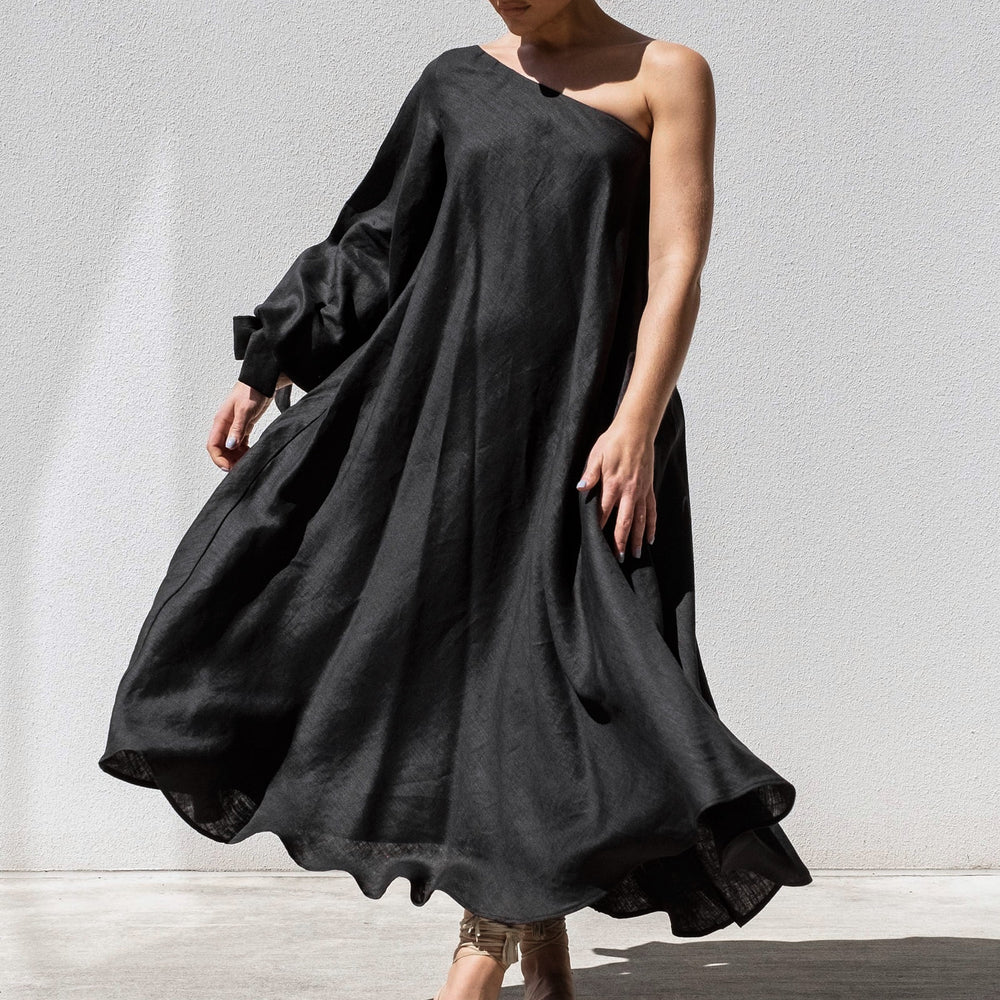 
                      
                        A person stands against a plain wall wearing the elegant Black Balloon Sleeve Swing Dress by Australian designer Isabella Longginou, featuring an asymmetrical off-the-shoulder design with flowing fabric. They are stepping forward while looking down and wearing open-toed heels.
                      
                    