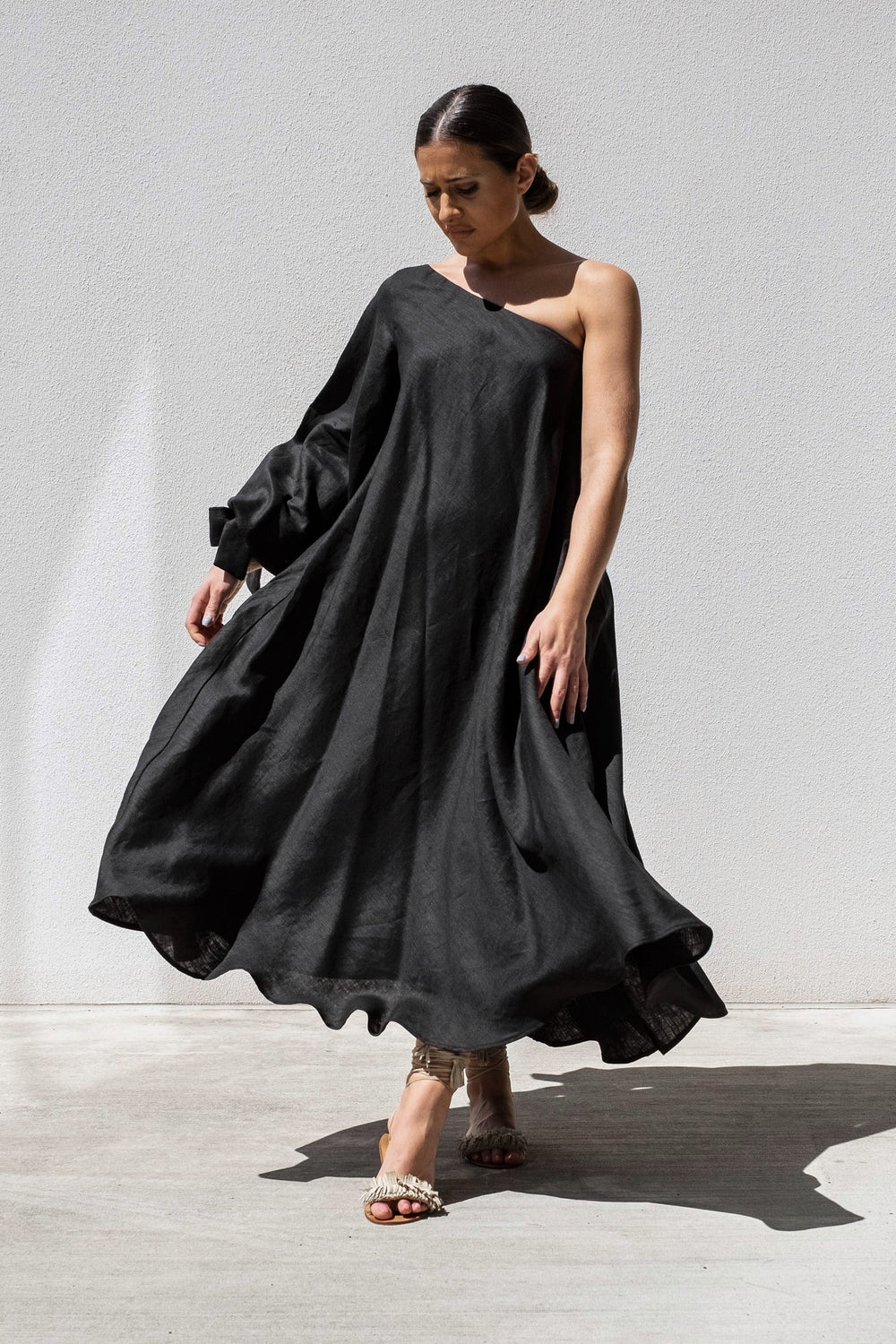 A person stands against a plain wall wearing the elegant Black Balloon Sleeve Swing Dress by Australian designer Isabella Longginou, featuring an asymmetrical off-the-shoulder design with flowing fabric. They are stepping forward while looking down and wearing open-toed heels.